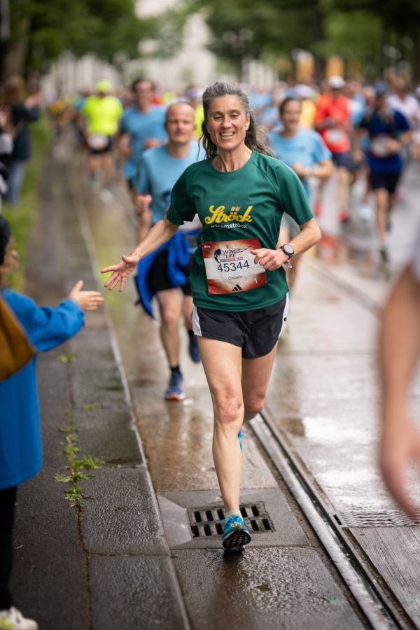 Eine Frau in grünem Hemd und Startnummer lächelt und gibt einem Kind ein High-Five, während sie auf einer nassen Straße in der Stadt mit anderen Läufern im Hintergrund an einem Marathon teilnimmt.