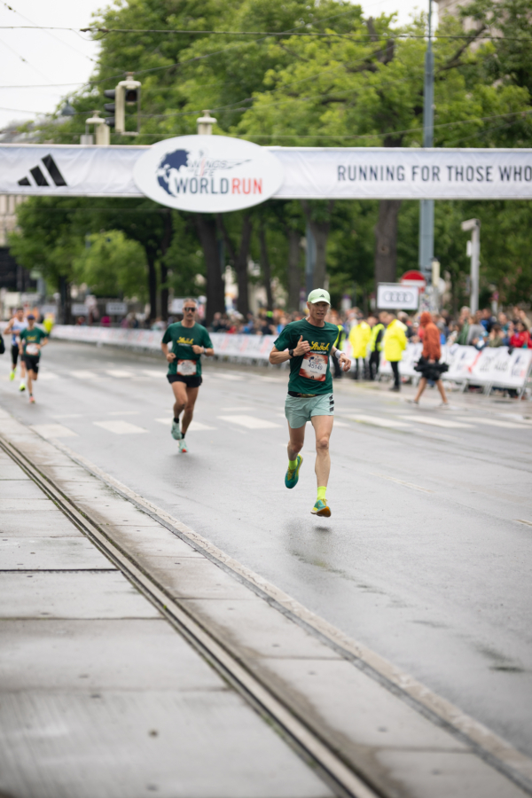 Läufer in grünen Hemden nehmen an einem regnerischen Tag an einem Stadtmarathon teil. Auf einem Banner darüber steht World Run und Running For Those Who Cant. Zuschauer säumen die Straße, und im Hintergrund sind Bäume und Gebäude zu sehen.