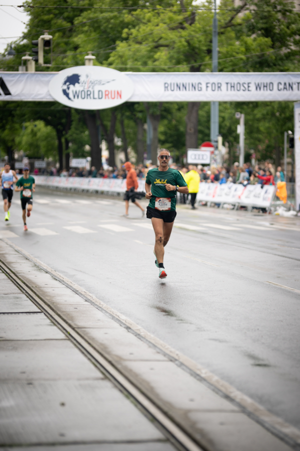 Ein Läufer in grünem Hemd und schwarzen Shorts nähert sich der Ziellinie eines Straßenlaufs, während die Zuschauer an einem bewölkten Tag hinter Absperrungen stehen und über ihnen ein großes World Run-Banner hängt.