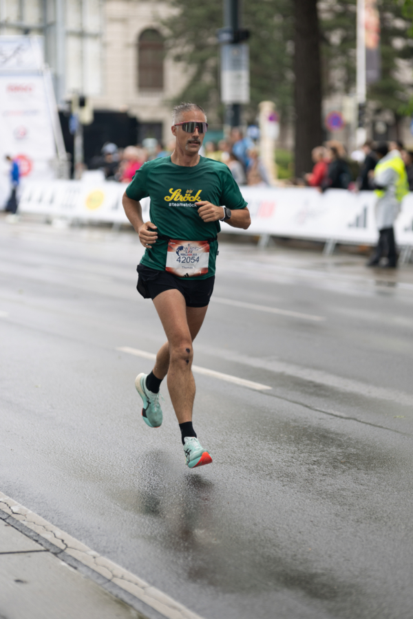 Ein Mann mit Sonnenbrille, grünem T-Shirt, schwarzen Shorts und Laufschuhen läuft während eines Rennens auf einer nassen Straße in der Stadt, mit einer Startnummer am Hemd und Zuschauern im Hintergrund.