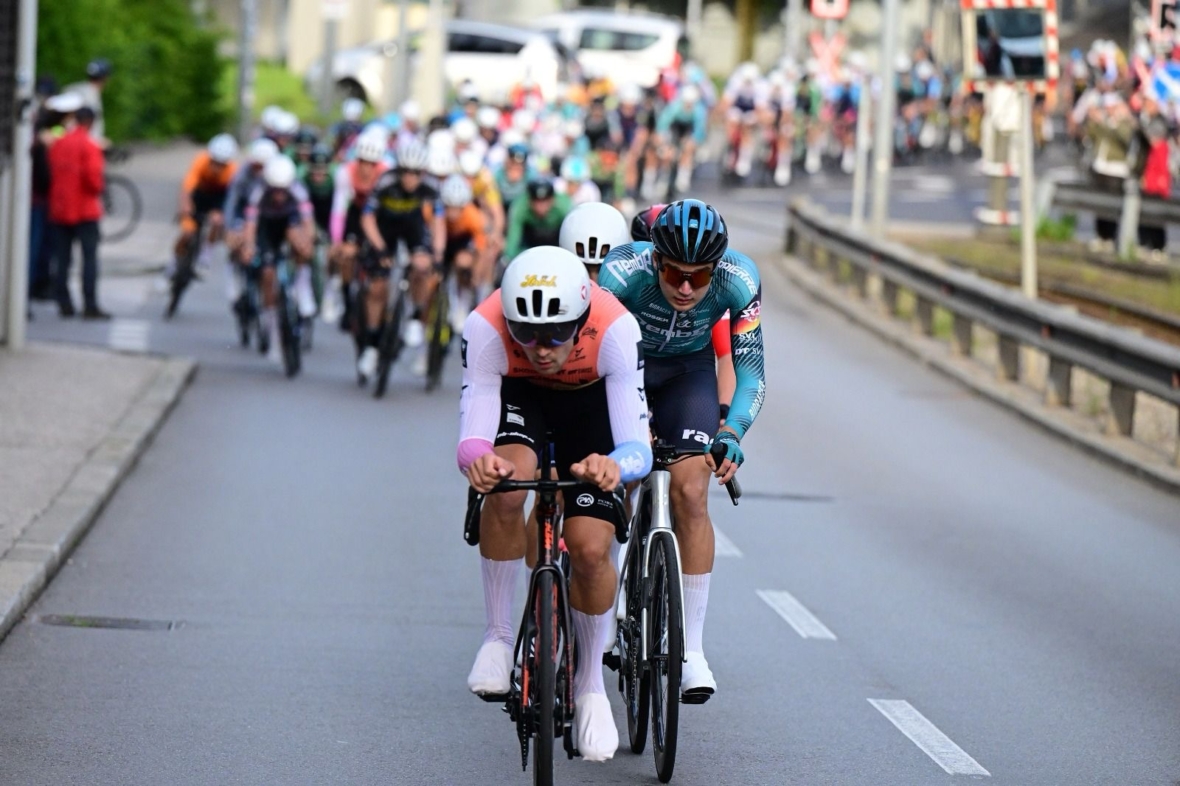 Eine Gruppe von Radfahrern nimmt an einem Straßenrennen teil, wobei zwei Fahrer das Feld auf einer Stadtstraße anführen. Im Hintergrund sind Zuschauer und Absperrungen zu sehen.