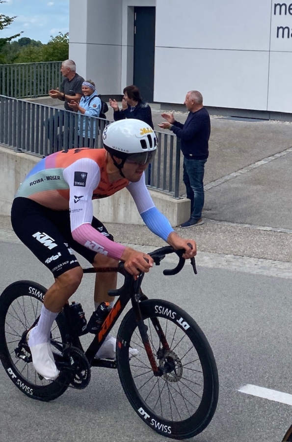 Ein Radfahrer in einem farbenfrohen Trikot und mit weißem Helm fährt während eines Rennens mit einem Rennrad bergauf, während eine kleine Gruppe von Menschen auf dem Bürgersteig hinter einem Geländer steht, klatscht und zusieht.