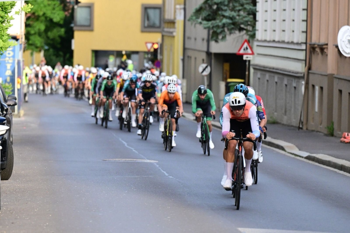 Eine Gruppe von Radfahrern rast eine Stadtstraße hinunter, die auf beiden Seiten von Gebäuden gesäumt ist. Die Fahrer tragen farbenfrohe Trikots und Helme, wobei der vorderste Radfahrer in einem rosa Oberteil die Gruppe anführt. Im Hintergrund sind Verkehrsschilder zu sehen.