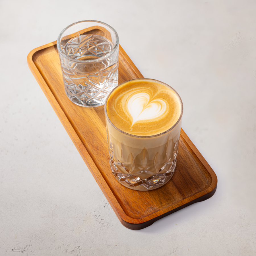 Ein Glas Milchkaffee mit herzförmiger Latte Art und ein Glas Wasser stehen auf einem rechteckigen Holztablett vor einem hellen Hintergrund.
