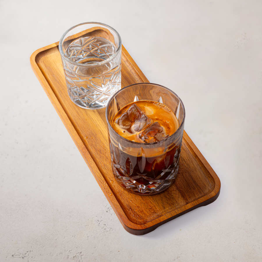 Ein Glas eisgekühlter Espresso und ein Glas Wasser stehen nebeneinander auf einem rechteckigen Holztablett, das auf einer hellen Fläche steht.