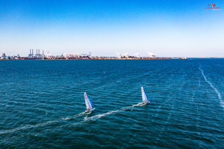 Zwei Segelboote fahren über tiefblaues Wasser auf eine entfernte Industrieküste mit Kränen und Gebäuden unter einem klaren, strahlend blauen Himmel zu.