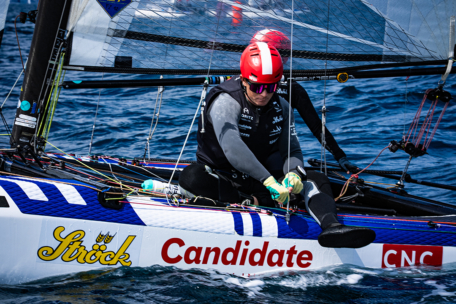 Ein Segler mit rotem Helm und Sonnenbrille richtet die Taue auf einem Rennsegelboot mit der Bezeichnung Strick Candidate CNC, das während einer Regatta über blaues Wasser gleitet.