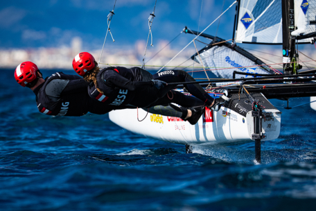 Zwei Segler mit roten Helmen und schwarzen Neoprenanzügen lehnen sich von einem Rennsegelboot aus waagerecht über das Wasser und balancieren bei einer Regatta um die Wette, während im unscharfen Hintergrund Stadtgebäude und Berge zu sehen sind.