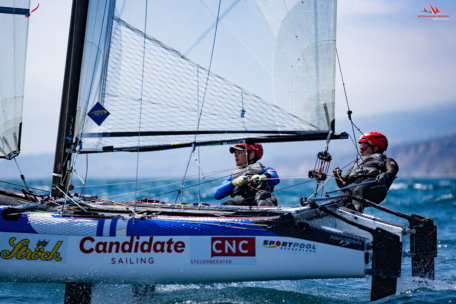Zwei Segler mit Helmen und Neoprenanzügen steuern ein schnelles Katamaran-Segelboot während einer Regatta und lehnen sich über das Wasser hinaus. Auf dem Boot sind verschiedene Sponsorenlogos zu sehen. Im Hintergrund sind blaue Meereswellen und eine ferne Küstenlinie zu sehen.
