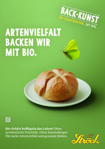 Ein rundes Brötchen liegt auf einem weißen Teller vor einem grünen Hintergrund. Ein grüner Schmetterling schwebt über dem Teller. Der Text lautet: "Artenvielfalt backen wir mit Bio." Branding: Ströck; wirbt für biologisches, biodiversitätsfreundliches Backen.
