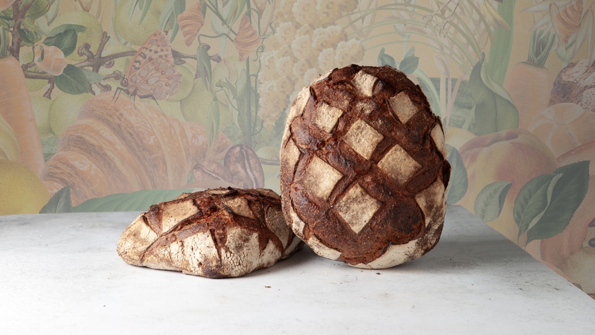 Zwei rustikale Brote aus Kittseer Bio-Holzofenbrot mit dunkler, kreuzschraffierter Kruste liegen auf einer hellen Fläche. Der Hintergrund zeigt eine farbenfrohe, botanische Illustration mit Früchten, Blättern und Blumen.
