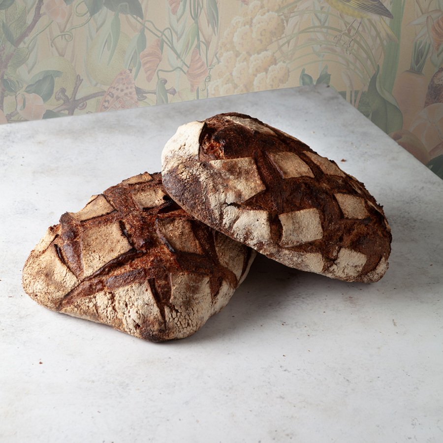 Zwei runde Laibe Kittseer Bio-Holzofenbrot mit dunkler, knuspriger Oberfläche, die ein Kreuzschraffurmuster aufweist, liegen auf einer hellen, strukturierten Oberfläche mit einer dezenten Blumentapete im Hintergrund.