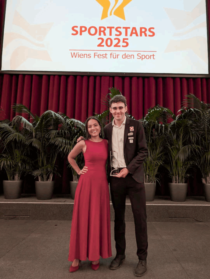 Zwei formal gekleidete Personen stehen lächelnd vor einer Bühne mit Topfpflanzen. Hinter ihnen ist eine große Leinwand mit der Aufschrift SPORTSTARS 2025 Wiens Fest für den Sport und roten Vorhängen zu sehen.
