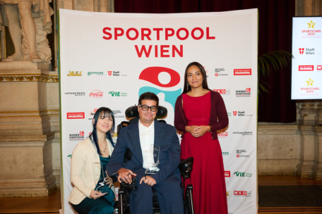 Drei Personen posieren bei einer Preisverleihung vor einem Sportpool Wien-Banner. Der Mann in der Mitte sitzt in einem Rollstuhl und hält einen Preis in der Hand, flankiert von zwei Frauen, eine in einem blaugrünen Kleid und eine in einem roten Kleid, die beide lächeln.