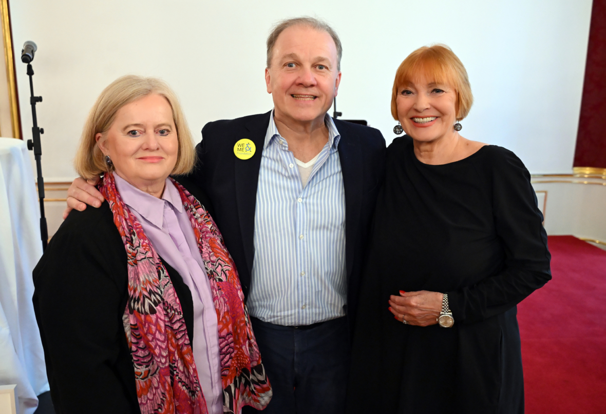 Drei Erwachsene stehen in einem Haus und lächeln in die Kamera. Die Frau links trägt einen rosafarbenen Schal und eine lavendelfarbene Bluse, der Mann in der Mitte einen Blazer und ein gestreiftes Hemd, und die Frau rechts trägt ein schwarzes Kleid.