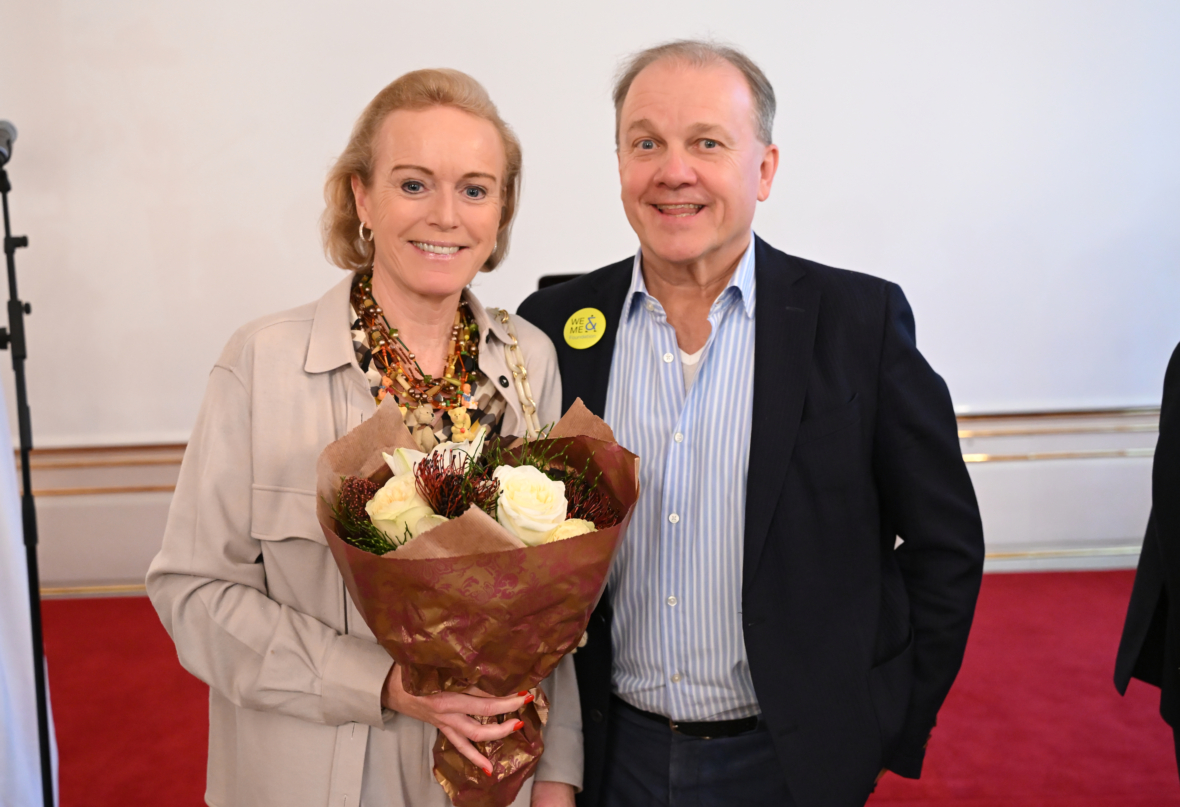 Eine lächelnde Frau, die einen Strauß weißer Blumen hält, steht neben einem lächelnden Mann. Beide sind halb-formal gekleidet, im Hintergrund liegen ein roter Teppich und eine helle Wand.