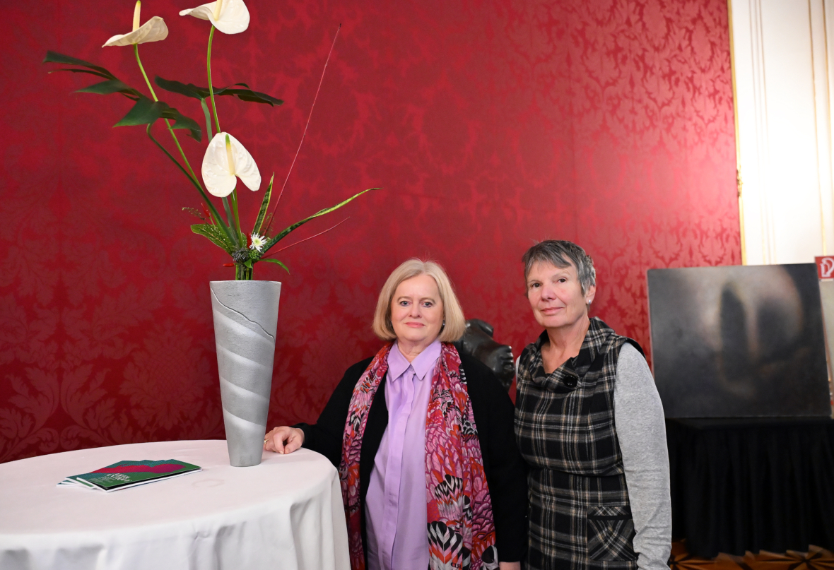 Zwei Frauen stehen neben einem runden Tisch mit einer hohen Vase mit weißen Blumen in einem Raum mit verzierten roten Tapeten. Auf dem Tisch liegt ein Büchlein, und im Hintergrund ist ein dunkles Kunstwerk zu sehen.