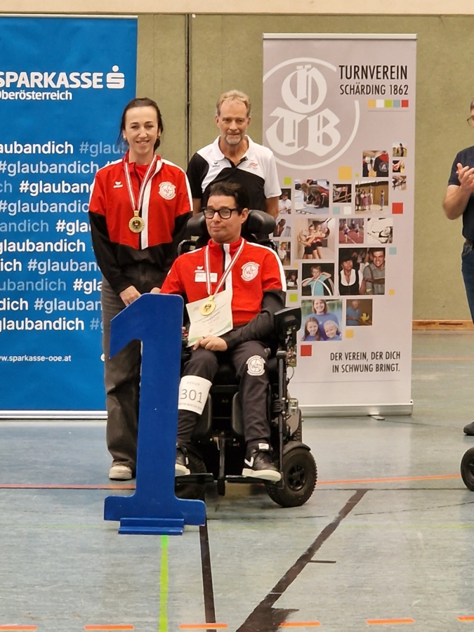 Two athletes in matching red and black uniforms, one standing and one in a wheelchair, both wearing gold medals, pose next to a blue 1 podium. A man stands behind them, smiling. Banners are visible in the background.