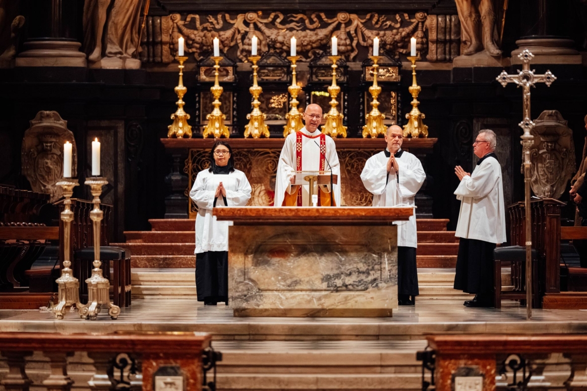 Vier Geistliche in weißen Gewändern stehen vor einem reich verzierten Altar mit hohen goldenen Kerzenleuchtern und religiösen Symbolen in einem großen Kirchenraum und bereiten sich auf eine religiöse Zeremonie vor.