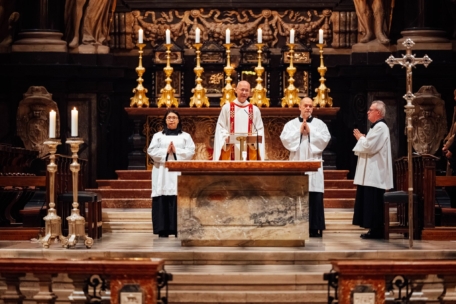Vier Geistliche in weißen Gewändern stehen vor einem reich verzierten Altar mit hohen goldenen Kerzenleuchtern und religiösen Symbolen in einem großen Kirchenraum und bereiten sich auf eine religiöse Zeremonie vor.