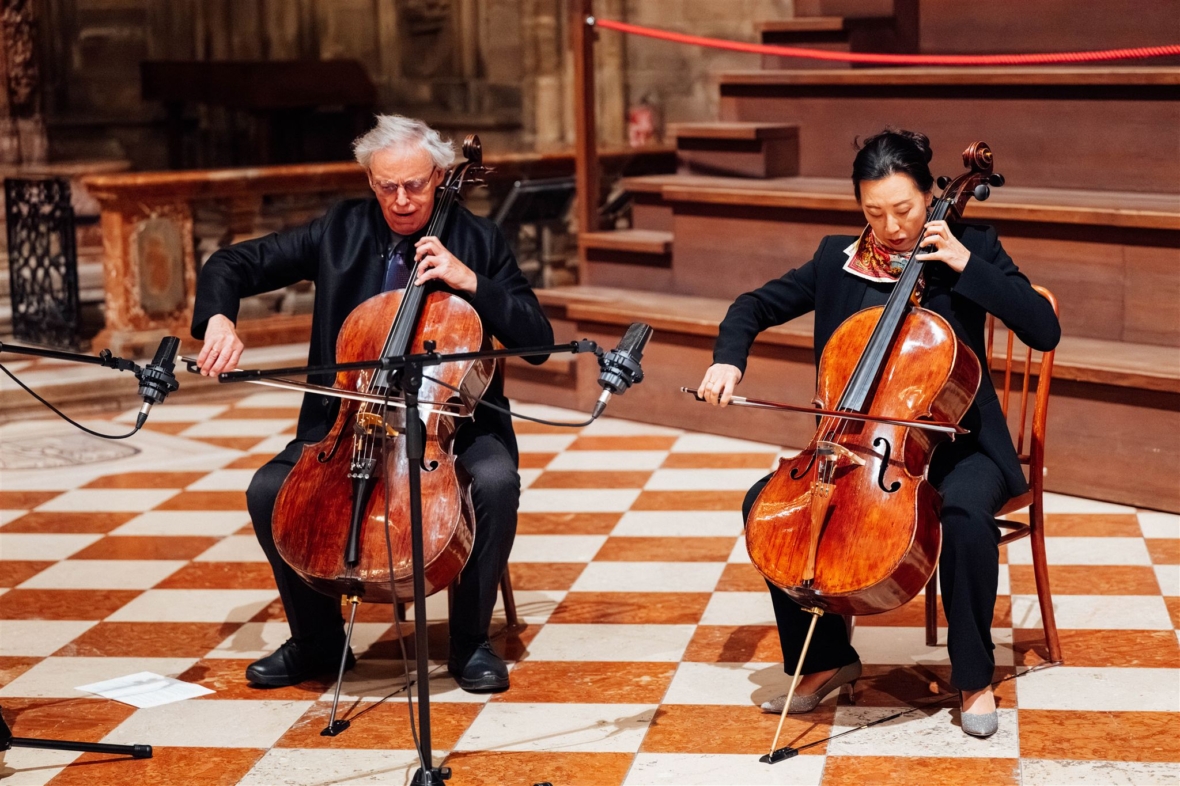Zwei Musiker sitzen auf Stühlen und spielen Celli auf einem karierten Fliesenboden in einem geräumigen, verschnörkelten Raum. Neben ihren Instrumenten sind Mikrofone aufgestellt, und beide sind auf ihr Spiel konzentriert.