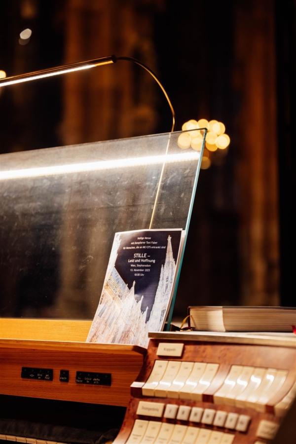 Eine Nahaufnahme eines Orgelspieltisches mit beschrifteten Tasten und Schaltern. Ein Musikprogramm mit der Abbildung einer Kathedrale liegt auf dem Ständer, beleuchtet von einer Schreibtischlampe in einer schwach beleuchteten Umgebung.