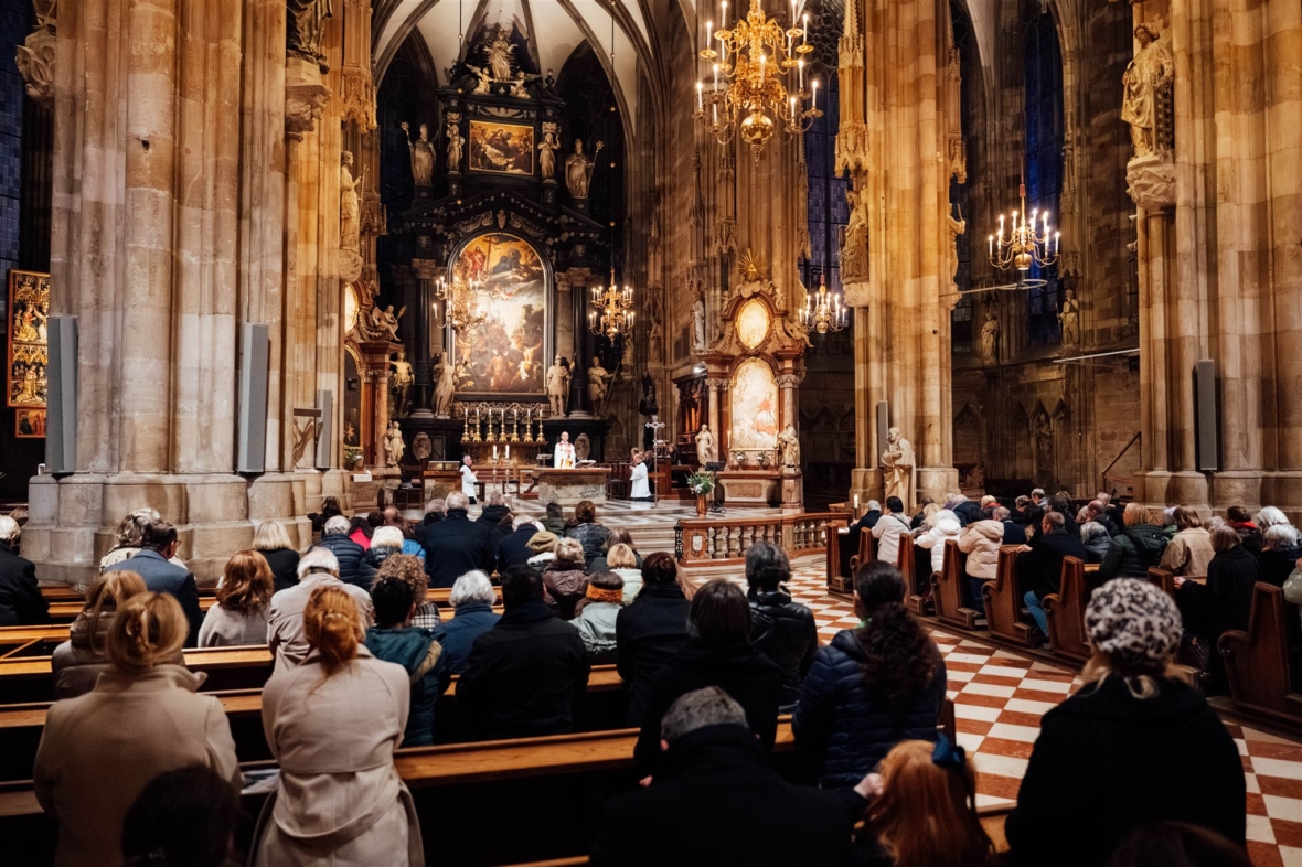 Die Menschen besuchen einen Gottesdienst in einer großen, reich verzierten Kathedrale mit hohen Bögen, Kronleuchtern und religiösen Kunstwerken. Die Gemeinde sitzt in Holzbänken gegenüber dem Altar, wo die Geistlichen den Gottesdienst abhalten.