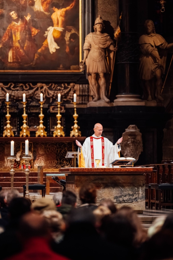 Ein Priester in weißem und rotem Gewand steht mit erhobenen Armen hinter einem Altar in einer großen Kirche, mit goldenen Kerzenleuchtern und religiösen Kunstwerken im Hintergrund. Im Vordergrund ist eine Gemeinde versammelt.