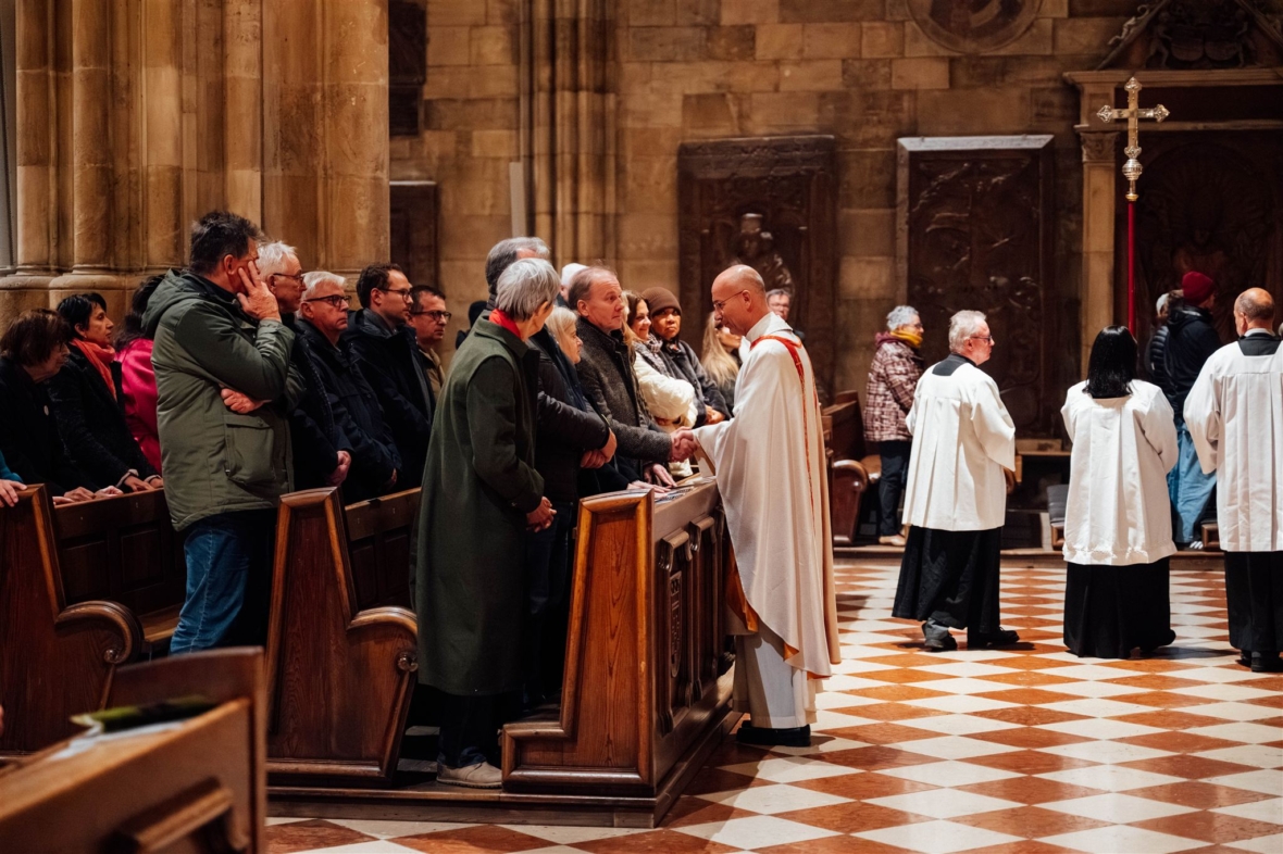 Ein Priester in weißem Gewand begrüßt die in den Kirchenbänken stehenden Gemeindemitglieder und schüttelt ihnen die Hand, während andere Geistliche in weißen Gewändern in der Nähe auf einem karierten Boden stehen.