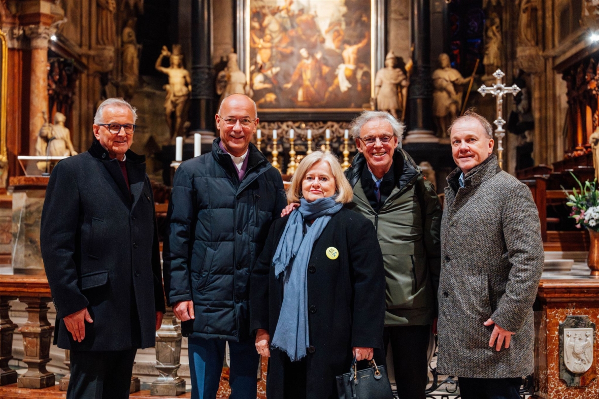 Fünf Personen stehen lächelnd in einer großen Kirche mit kunstvollen Verzierungen, Statuen und einem großen Gemälde im Hintergrund. Vier Männer tragen Mäntel und eine Frau steht in der Mitte mit einem Schal und einer Tasche.