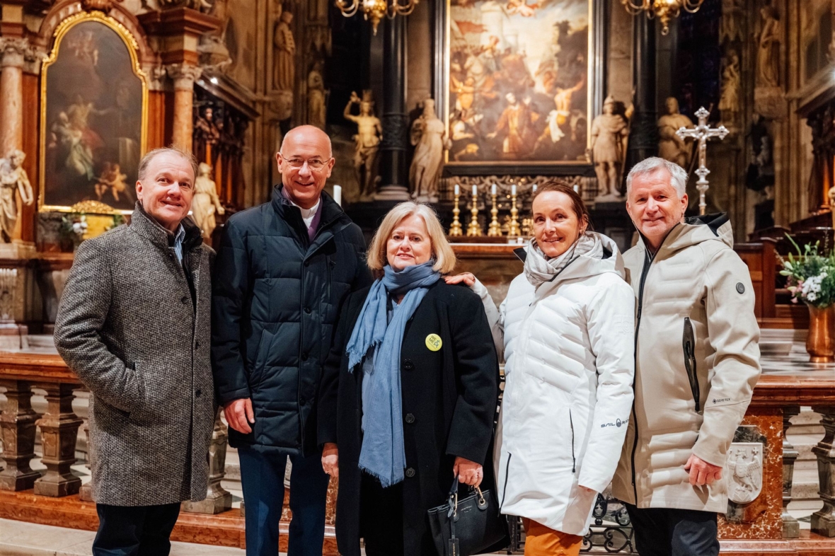 Fünf Erwachsene stehen in einer verzierten Kirche, gekleidet in Wintermäntel und Schals, und lächeln in die Kamera. Im Hintergrund sind religiöse Kunstwerke, Statuen und ein geschmückter Altar zu sehen.