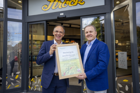 Thomas Pucharski und Gerhard Ströck lächeln und halten eine gerahmte Urkunde in der Hand, während sie vor einem Ladeneingang stehen, über dessen Tür ein Schild mit der Aufschrift "Ströck" hängt. Im Hintergrund sind Regale mit Produkten und eine Glastür zu sehen.