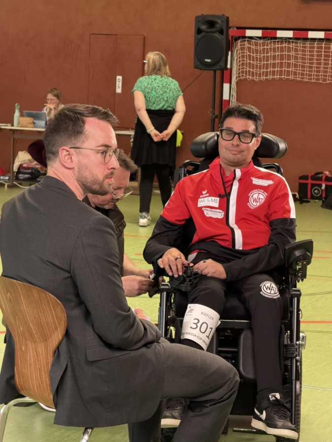 Ein Mann im Anzug sitzt einem anderen Mann im Rollstuhl gegenüber, der eine rote Sportjacke mit der Nummer 301 trägt. Sie befinden sich in einer Turnhalle mit Menschen und Sportgeräten im Hintergrund.