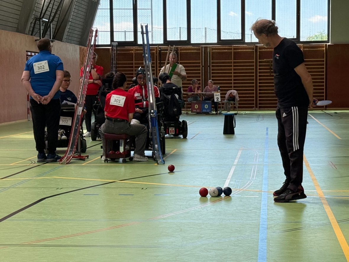 Boccia-Spiel in der Halle. Mehrere Spieler in Rollstühlen, die nummerierte Lätzchen tragen, werfen die Kugeln über Rampen. Ein stehender Schiedsrichter beobachtet das Spielfeld, auf dem rote und blaue Kugeln in der Nähe der weißen Zielkugel positioniert sind.