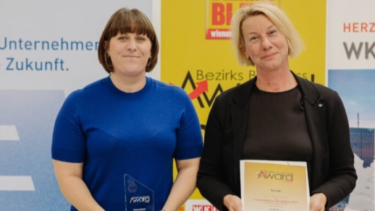Stephanie Dürrstein und Andrea Unger-Posch stand side by side at an award event. One holds a glass award, the other holds a certificate. Both are smiling. There are banners and signs with German text in the background.