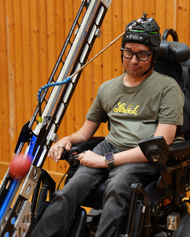 Ein Mann, der einen am Kopf befestigten Zeiger trägt und in einem Elektrorollstuhl sitzt, benutzt diesen, um einen roten Ball von einer daran angebrachten Rampe abzuschießen.