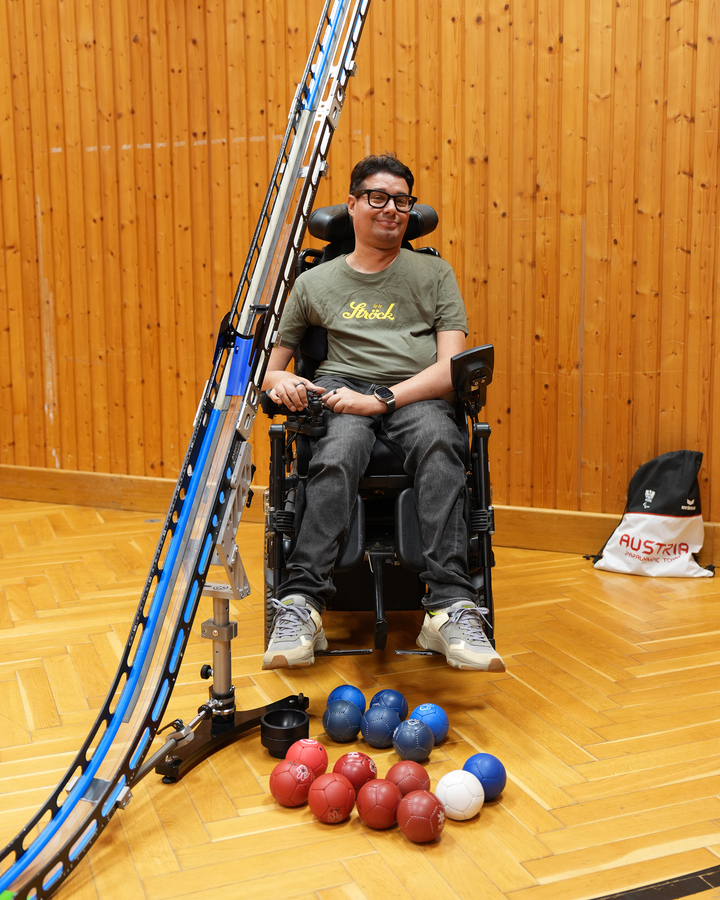 Ein Boccia-Spieler in einem Elektrorollstuhl sitzt neben einer Rampe, auf deren Boden rote, blaue und weiße Kugeln liegen.