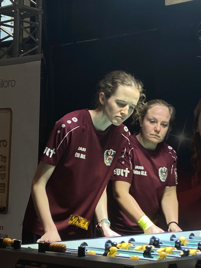 Zwei Frauen in passenden kastanienbraunen Hemden spielen zusammen Tischfußball und konzentrieren sich auf das Spiel. Die Szene befindet sich in einem Innenraum mit heller Beleuchtung über dem Tischfußballtisch.