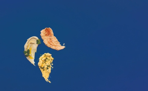 Drei verschiedene Aufstriche - ein beiger mit Petersilie, ein orangefarbener mit Paprika und ein gelber mit Schnittlauch - sind in einer diagonalen Linie vor einem einfarbig blauen Hintergrund angeordnet.