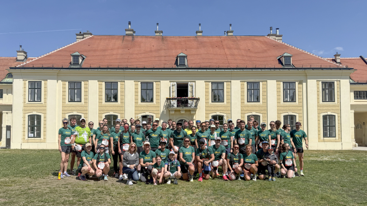 Eine große Gruppe von Menschen in passenden grünen Hemden posiert für ein Foto auf dem Rasen vor einem gelben Gebäude mit rotem Dach, unter einem klaren blauen Himmel.