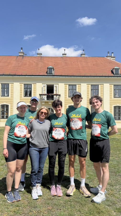 Sechs Personen stehen an einem sonnigen Tag lächelnd vor einem großen gelben Gebäude mit rotem Dach. Vier von ihnen tragen passende grüne Laufshirts und Startnummern, einer trägt Freizeitkleidung und einer eine weiße Mütze und Sonnenbrille.