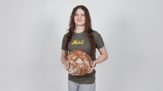 Eine Person mit langen braunen Haaren, die ein grünes T-Shirt und eine graue Hose trägt, steht vor einem schlichten hellen Hintergrund, hält einen großen runden Laib rustikalen Brotes in der Hand und lächelt sanft.
