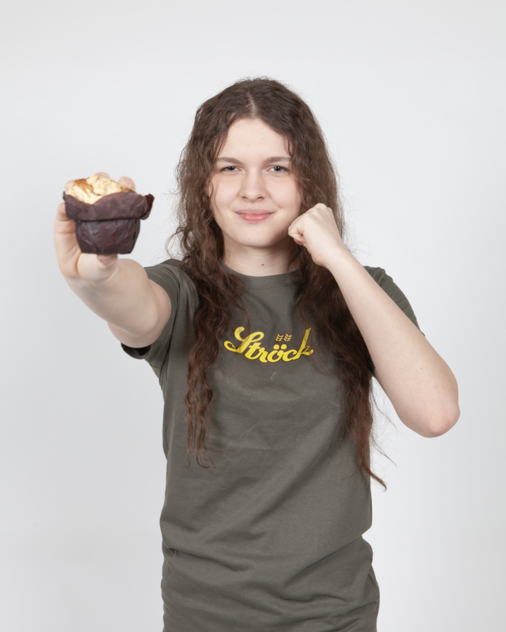 Eine junge Person mit langen braunen Haaren, die ein grünes Ströck-T-Shirt trägt, lächelt und hält mit einer Hand einen Muffin in die Kamera, während sie mit der anderen Hand eine Faust macht. Der Hintergrund ist einfarbig weiß.
