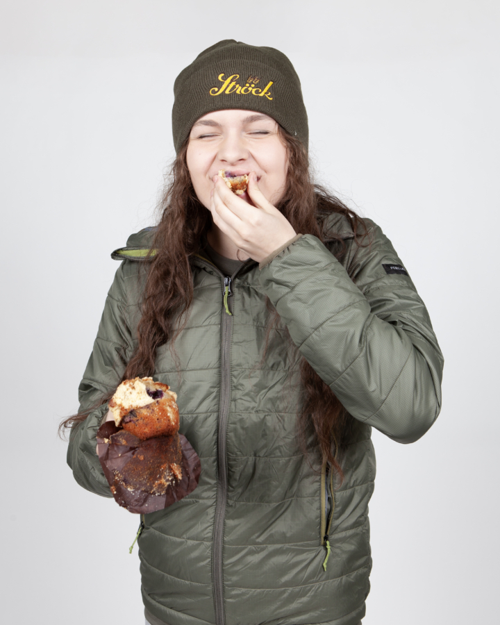 Eine Person in grüner Jacke und Mütze lächelt mit geschlossenen Augen, während sie einen Muffin isst und ein weiteres Stück in der Hand hält, vor einem schlichten hellen Hintergrund.