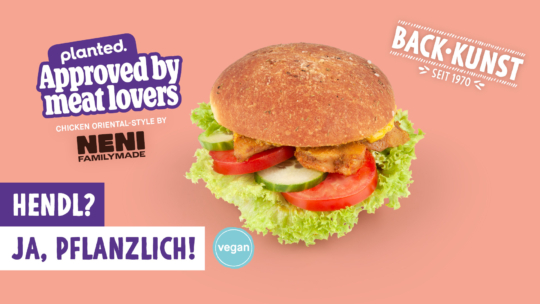 Ein veganes Hühnchensandwich mit Salat, Tomate und Gurke auf einem entkernten Brötchen wird mit einem Text gezeigt, der für Protein-Power auf pflanzlicher Basis wirbt, mit Aufschriften wie Approved by meat lovers und Ja, pflanzlich! auf einem rosa Hintergrund.