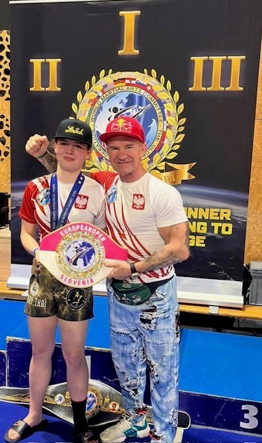 Zwei Personen stehen lächelnd vor einem Banner für eine Kampfsportmeisterschaft. Der eine, in kurzen Hosen und mit einer Medaille, hält eine Auszeichnung "European Open Slovenia" in der Hand. Der andere, mit rot-weißem Hemd und Hut, hat den Arm um den Athleten gelegt.