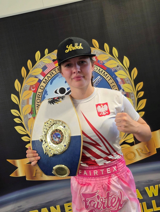 Eine Kampfsportlerin mit Mütze und Sportbekleidung hält einen Meistergürtel in der Hand und ballt vor einer Kulisse des World Martial Arts Committee die Faust.