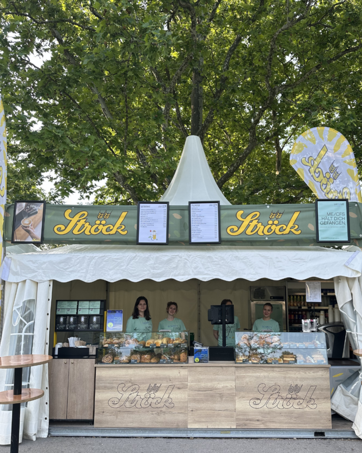 Ein Bäckereistand mit der Aufschrift "Ströck" mit drei Angestellten hinter der Theke, die verschiedene Backwaren anbieten. Der Stand befindet sich im Freien, unter einem großen Baum, mit zwei hohen Schildern und einem weißen Vordach.