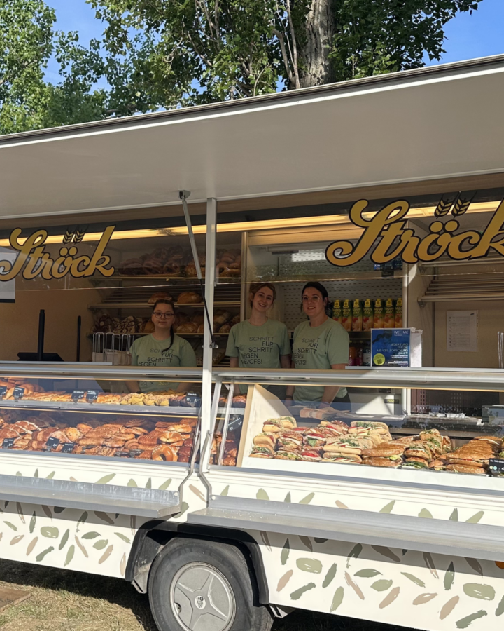 Drei Personen stehen hinter dem Tresen eines Bäckereiwagens von Ströck, in dem Regale mit Gebäck, belegten Brötchen und Getränken zu sehen sind. Der Wagen ist an einem sonnigen Tag im Freien geparkt, mit Bäumen im Hintergrund.