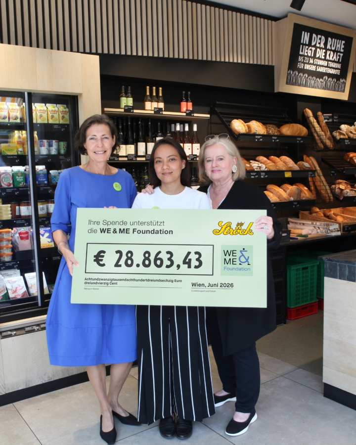 Drei Frauen stehen in einer Bäckerei und halten einen großen Scheck über 28.863,43 €, ausgestellt auf die WE & ME Foundation. Im Hintergrund sind Brotlaibe und Regale mit Lebensmitteln zu sehen.